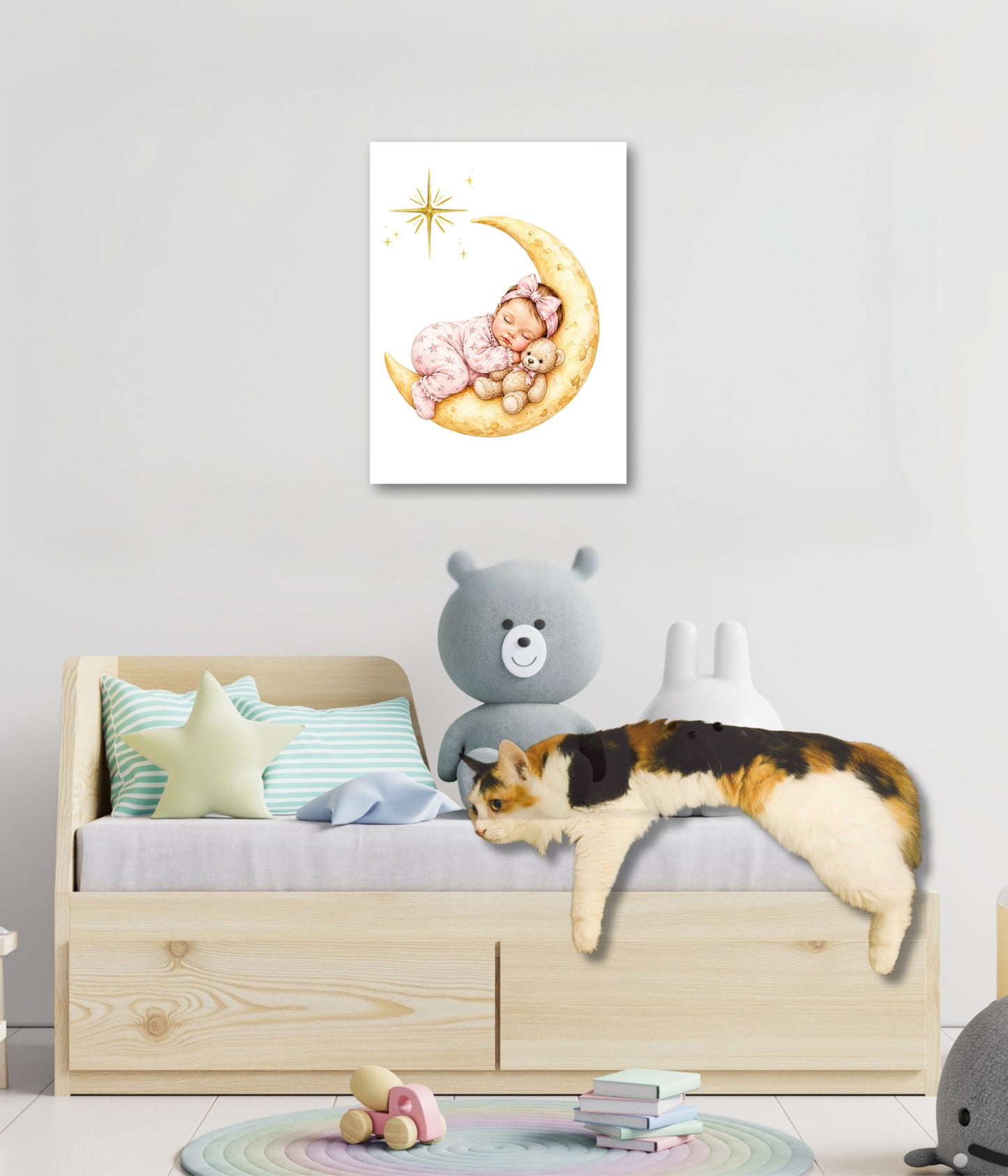 A children's bedroom scene with a light wood bed, pastel pillows, a gray teddy bear, and a real cat lying across the bed. Above the bed hangs a framed watercolor-style print showing a baby sleeping on a crescent moon with a star.
