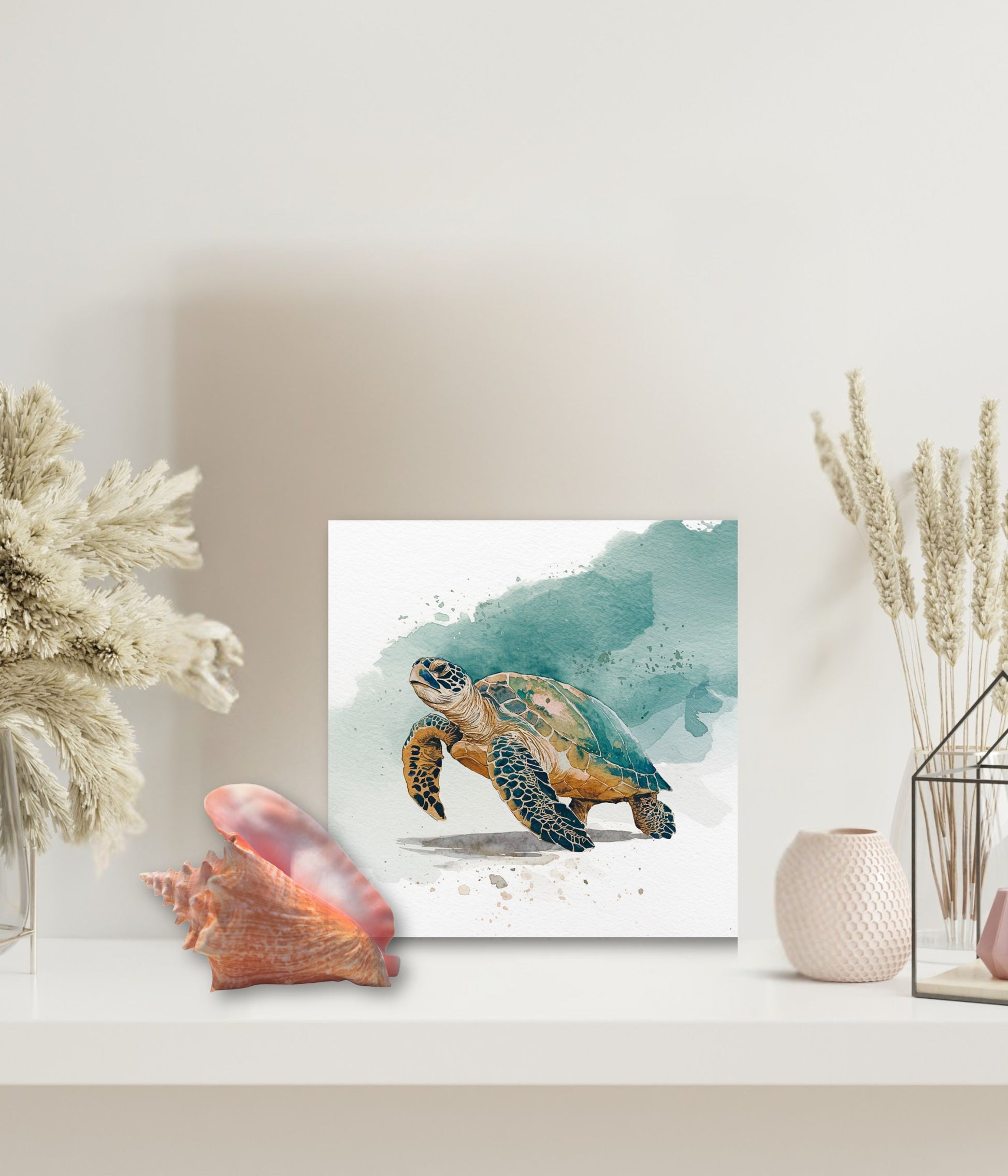 A square watercolor-style art print of a sea turtle displayed on a white shelf, surrounded by decorative props including dried grasses, a pink shell, and a textured vase in a bright, minimal interior setting.