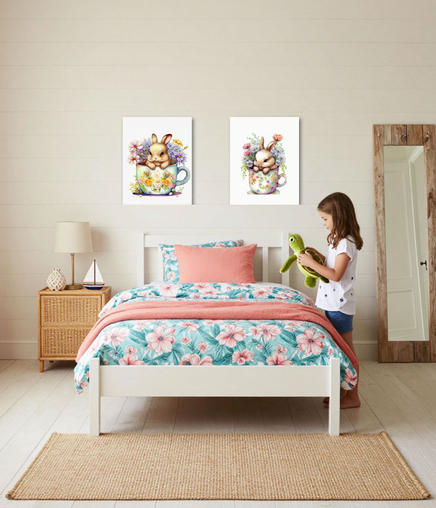 A child's bedroom scene with a white bed dressed in a pink and teal floral print, coral throw blanket, and matching pillow. Two framed bunny artwork prints hang above the bed.