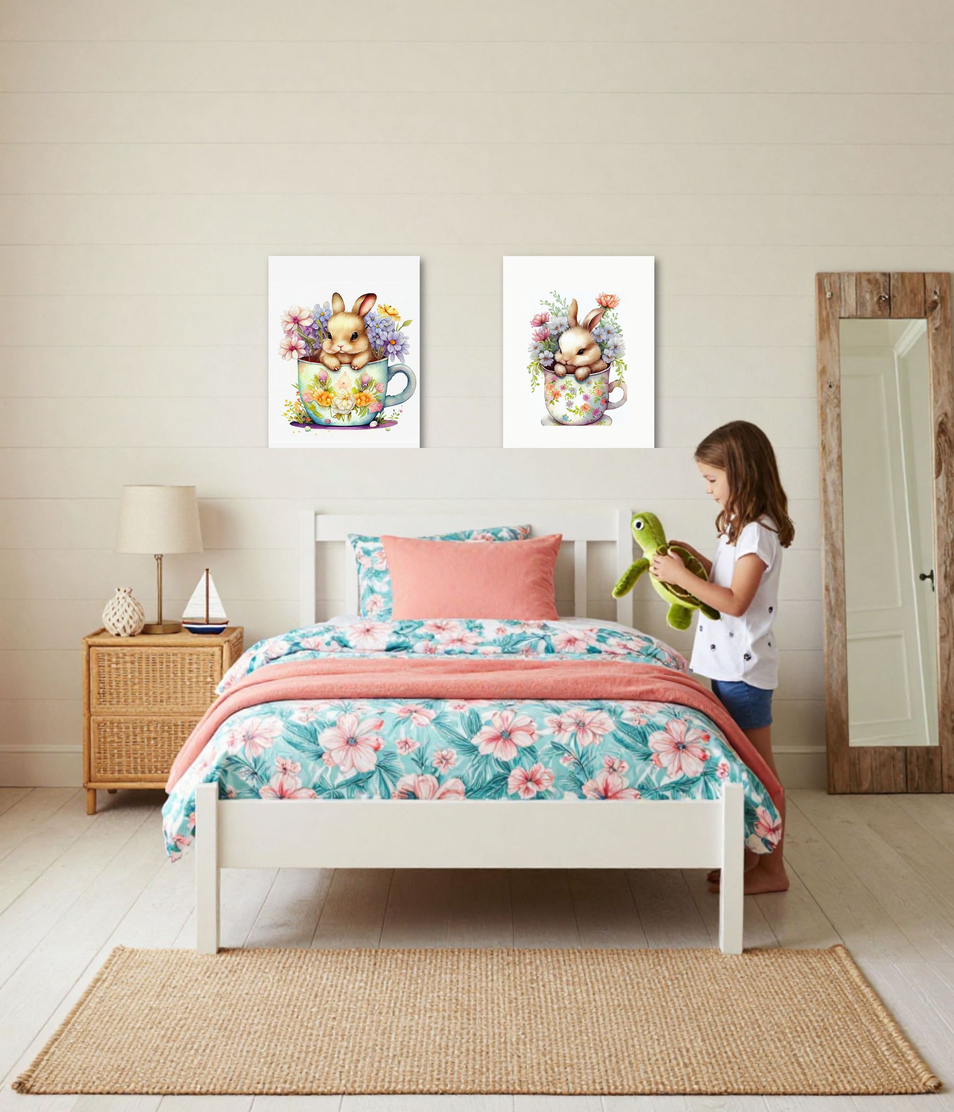 A child's bedroom scene with a white bed dressed in a pink and teal floral print, coral throw blanket, and matching pillow. Two framed bunny artwork prints hang above the bed.