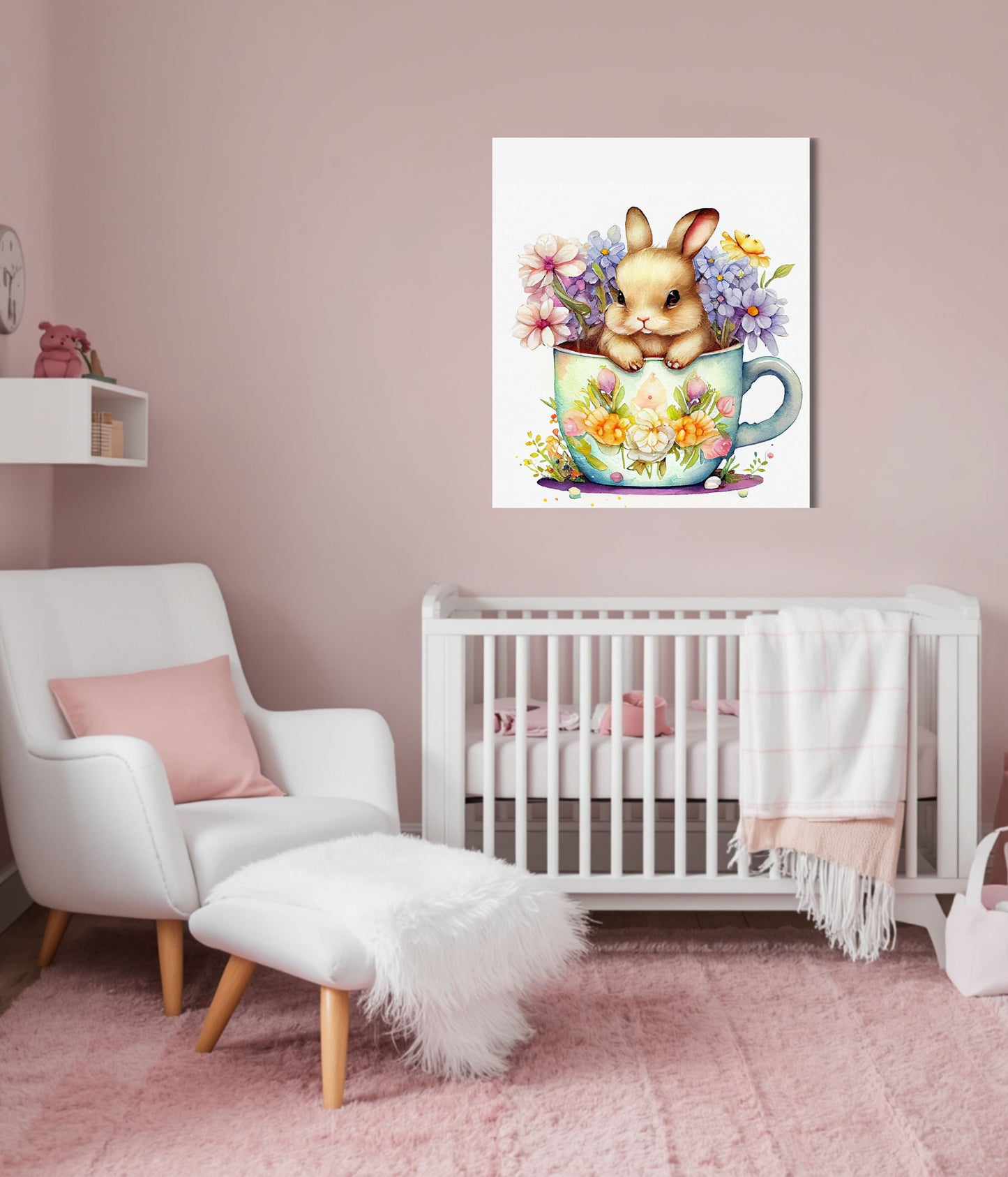 A framed wall art print showing a small bunny sitting in a floral teacup, displayed above a white crib in a soft pink nursery with a white chair and pink rug.
